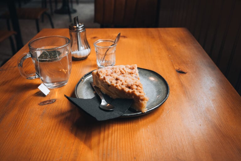 A slice of apple strudel served on a round plate beside a steaming cup of tea at Au Fond de la Cour café in Obernai. The dessert is lightly dusted with powdered sugar and sits on a small outdoor café table.