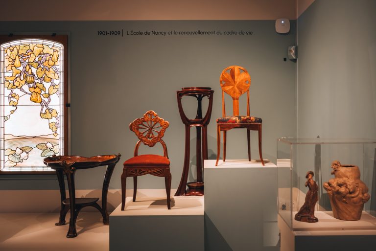 This is an Art Nouveau Exhibit at the Fine Arts Museum in Nancy. There are two chairs, a small table, a large stand, and a stained-glass window.