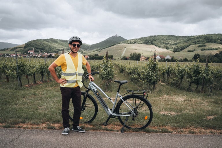 This is Cedric Schuhler standing next to my (Jen Ciesielski) bike while giving me a bike tour. He is wearing a helmet and a reflective vest. Behind him are vineyards and rolling mountains.