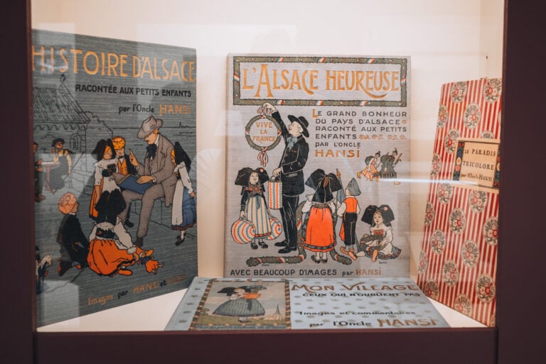 Display of children’s books illustrated by Hansi inside The Village Hansi museum showing colorful vintage style illustrations of Alsatian life. The exhibit highlights regional art and culture.