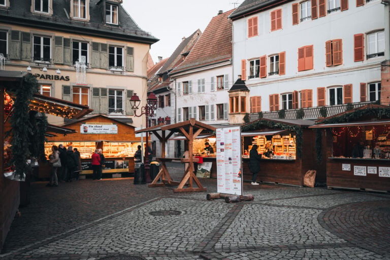Christmas market stalls decorated with wreaths and lights in s in Place Saint-Thiébaut in Thann. People are walking around and shopping. It's not busy or overcrowded.