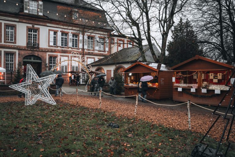 This is the Christmas market at the Parc du Musée in Wissembourg. There are wooden chalets and Christmas decorations. People are walking around.