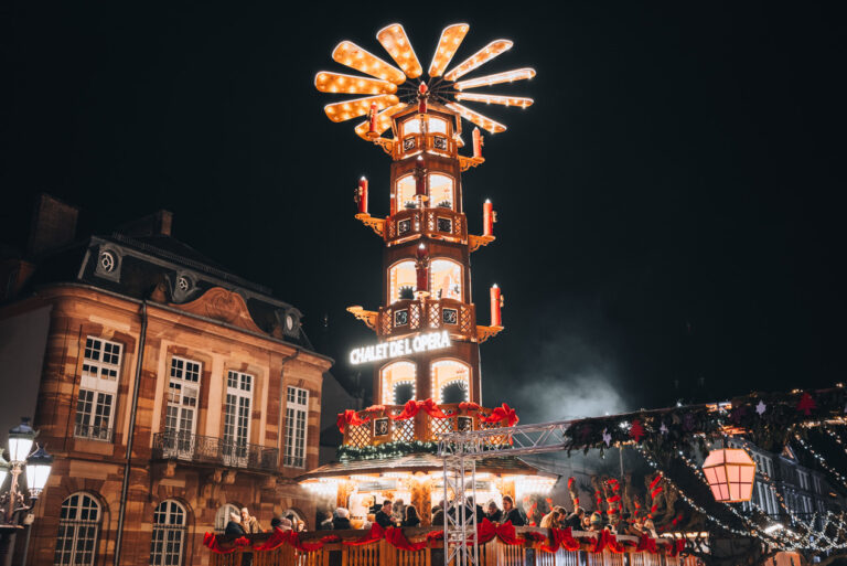 Wooden chalet with a tower and a terrace at the Christmas market in Place Broglie in Strasbourg at night. The lights shine bright and people are drinking vin chaud on the upper terrace.
