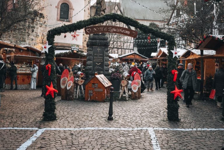This is the Christmas market in Place du Marché Saules in Eguisheim. There is a sign that says Noel à Eguisheim and it's under and arch decorated with wreaths. Behind the sign there are wooden Christmas stalls.