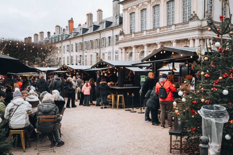 This is the Christmas market in front of the Palais du Gouvernement in Nancy. There are huts selling food and people are eating, drinking, and walking around.