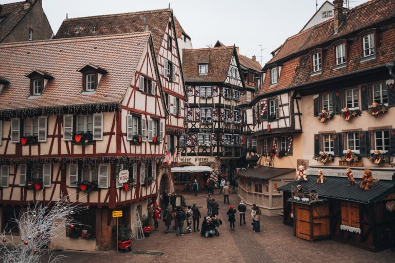 This is Rue des Marchands the Colmar Christmas Market. There are several half-timbered houses decorated with lights and ornaments. People are walking around.