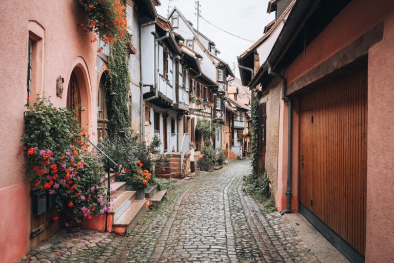 Brightly colored half timbered houses lining Rue du Rempart in Eguisheim with flower boxes, cobblestone streets, and a circular village layout.