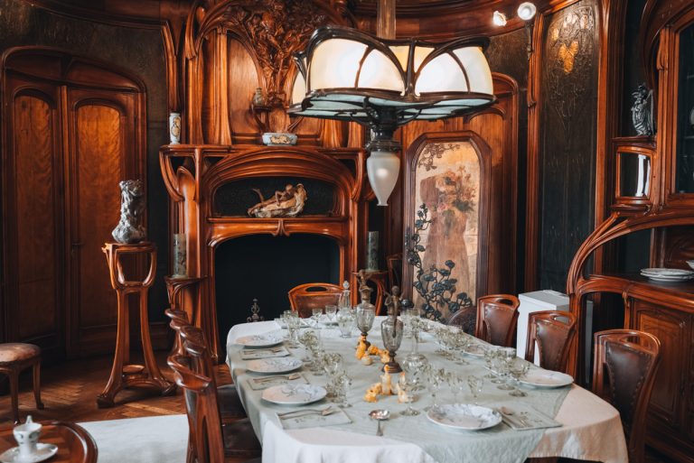 This is dining room set with Art Nouveau furniture at the Ecole de Nancy Museum. There is a table surrounded by chairs and several cabinets behind the table.
