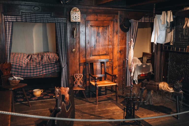 This is a photo of one of the bedrooms at the Musée Alsacien in Strasbourg. The walls and floor are made of wood. There are two beds, a wooden chair, and a rocking horse. In the corner clothes are hanging from a wood burning heater.