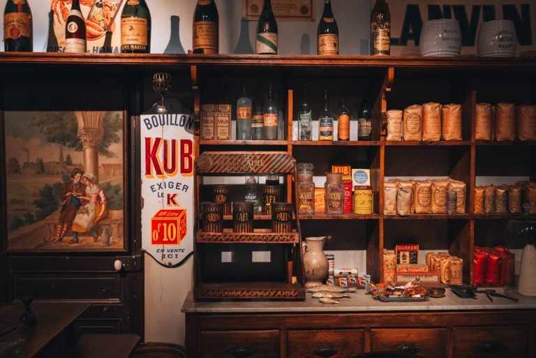 This is an exhibit at the Museum of Burgundian Life in Dijon. It shows in the side of a shop from the 19th century. There are shelves with various items like pop, wine, and other baking items. 