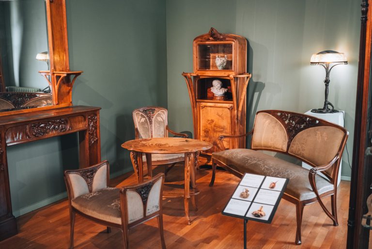 This is an furniture exhibit at the École de Nancy Museum. There is a table surrounded by chairs and behind it is a cabinet with two statues and a lamp.