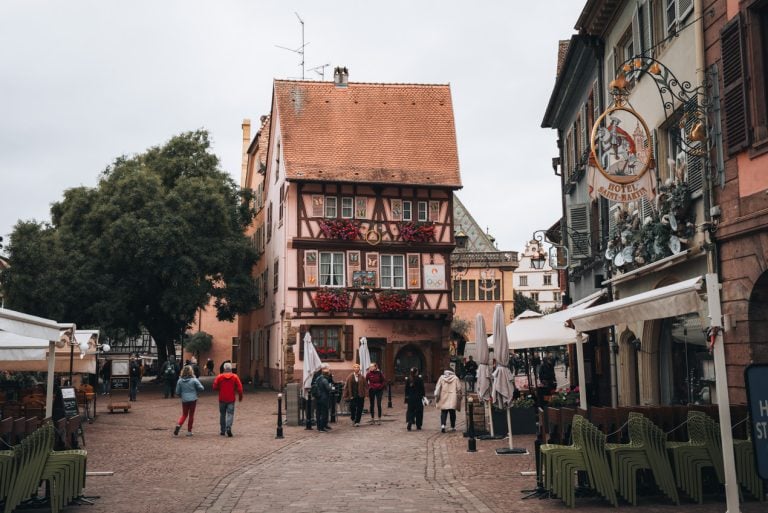 This is Grand Rue in the Old Town of Colmar. There is a colorful half-timbered house along a cobbled street and people are walking around. on the right there is a sign for the Hotel Saint Martin.