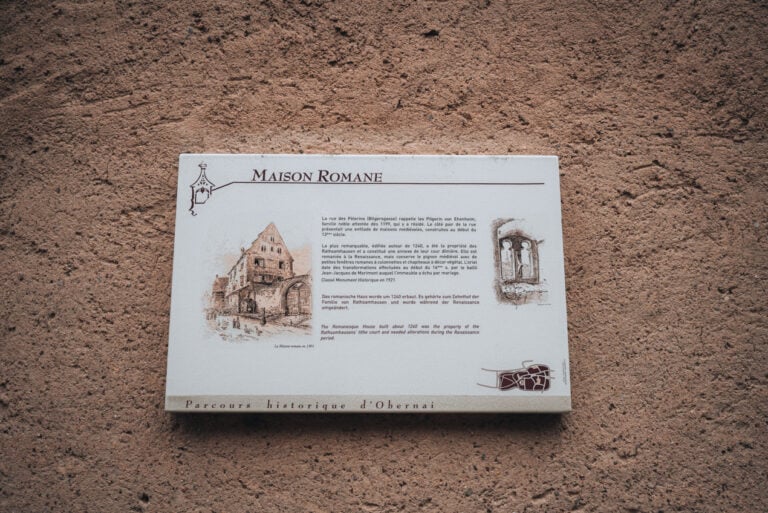 Wooden information board describing the history of the Romanesque House in Obernai with paragraphs of text written in French German and English. The sign stands along the Old Town Circuit where visitors stop to learn about historic buildings around the town.