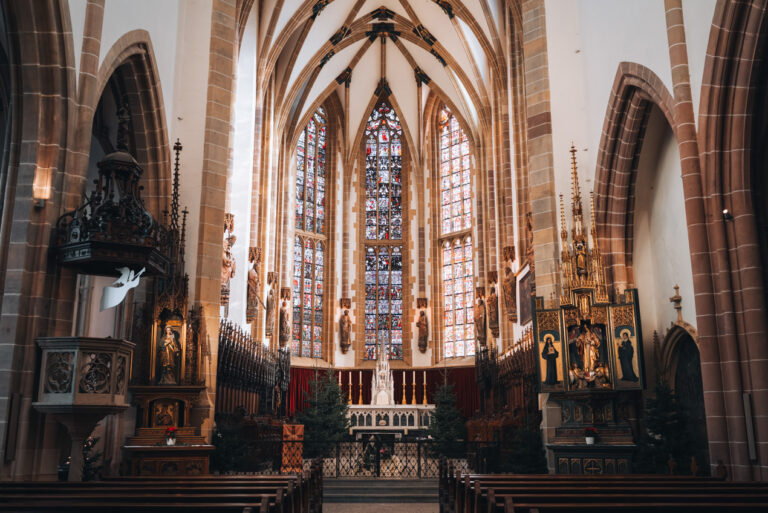 A row leading to an altar that is surrounded by stained glass windows and 12 statues of the Apostles inside Saint Theobald’s Church in Thann.