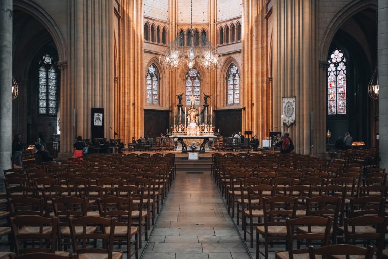 This is the inside of the 
Cathédrale Saint-Bénigne de Dijon. There is an aisle leading to the altar. It's surrounded by rows of wooden chairs. The altar is surrounded by three stained-glass windows and there is a chandelier that is shinning light on it.