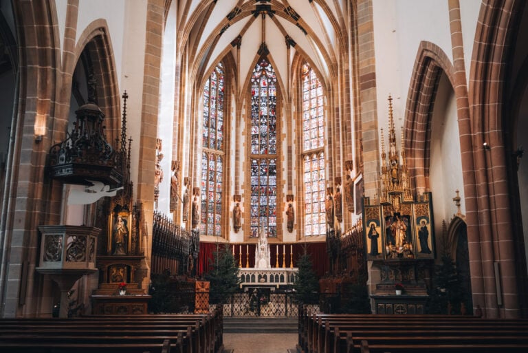 An aisle surrounded by rows of wooden chairs lead to the altar inside the Collegiate Church of Saint Thiébaut in Thann. Christmas trees and stained-glass window surround the lit altar to give it that Christmas time feel.