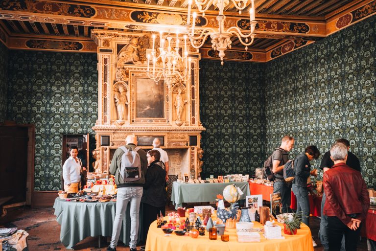 This is the inside of Hotel de Vogue. It's a small room that is hosting a craft market. There are tables with various items and people shopping. The room is decorated with colorful wallpaper, a stone fireplace, and a chandelier. 