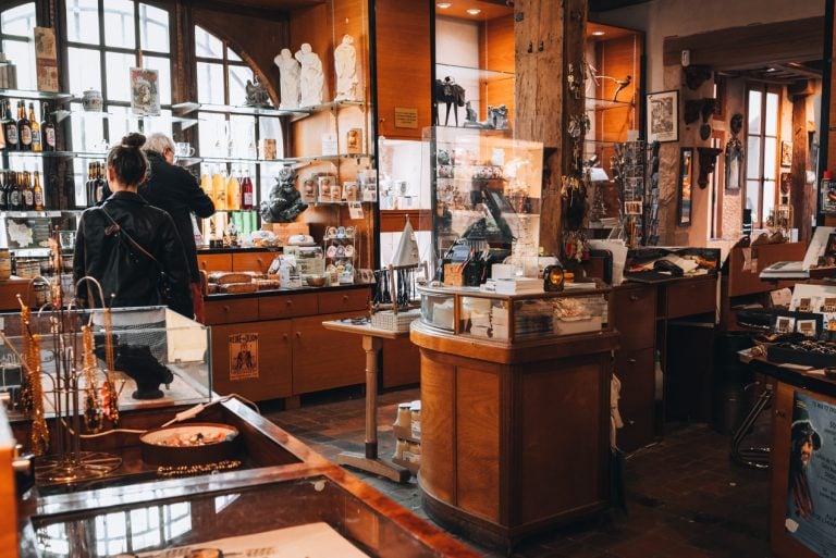 This is the interior of the boutique section at the Maison Millière in Dijon. There are selves and stands all around the store and they all have different items like books, jewelry, etc. There are also people shopping. 