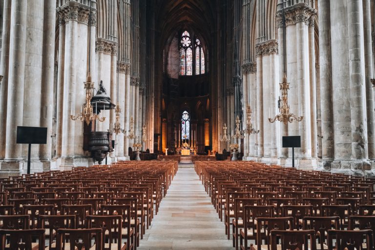 This is the inside of the Reims Cathedral. There is a aisle leading to the altar and it's surrounded rows of wooden chairs. The atlar is surrounded by stained glass windows.