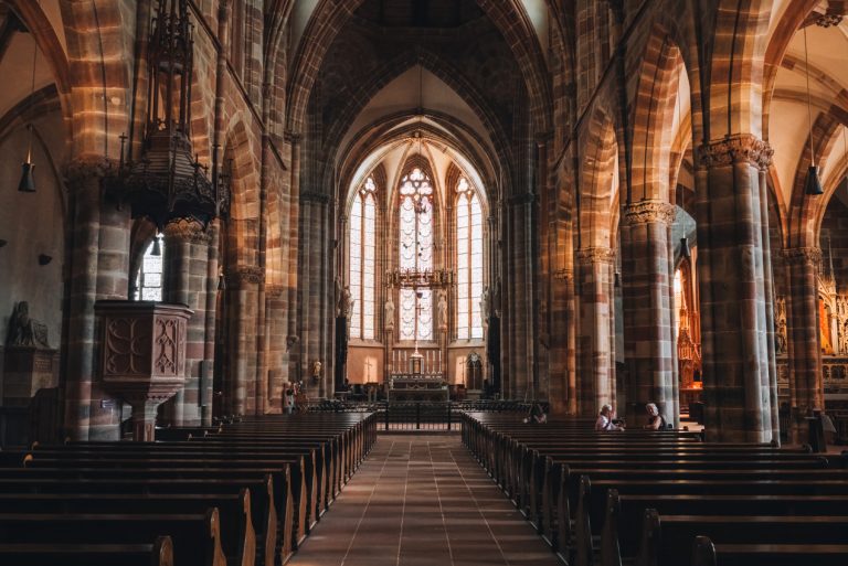 This is the inside of the Saint Peter and Saint Paul Abbey in Wissembourg. There is a row lined with pews leading to the altar. The altar is surrounded by stained glass windows.