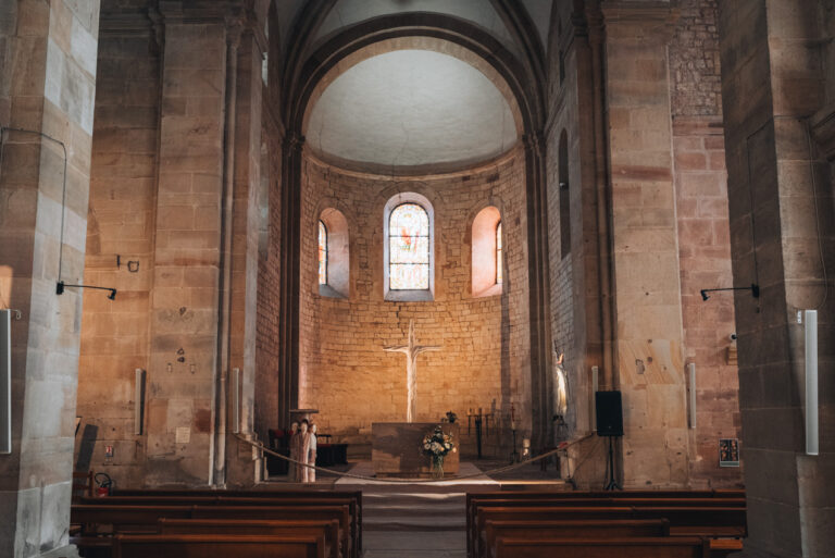 nside the Saints Peter and Paul Church in Rosheim. A narrow aisle leads to the altar where there are three stained glass windows and a cross with Jesus.