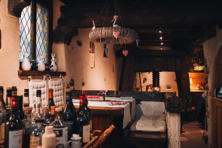 Interior of Caveau du Vigneron in Turckheim with wooden tables and seats, and bottles displayed along rustic stone walls.