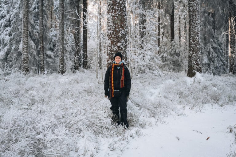 Jen standing in the snow at Champ du Feu surrounded by pine trees and a winter landscape in the Vosges Mountains. A snowy day trip from the city is one of the outdoorsy things to do in December.
