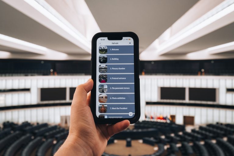 This is a photo of Jen holding her phone in the Hemicycle room of the European Parliament in Strasbourg. It shows the audio guided tour she is following and what she is listening to.