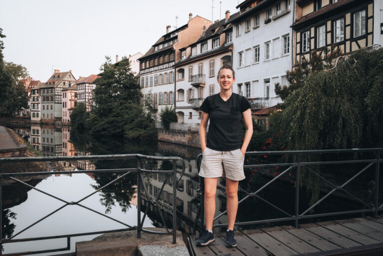 Jen standing on the Pont du Faisan bridge in Strasbourg’s Petite France district, with half-timbered houses and a calm canal in the background on a clear day.