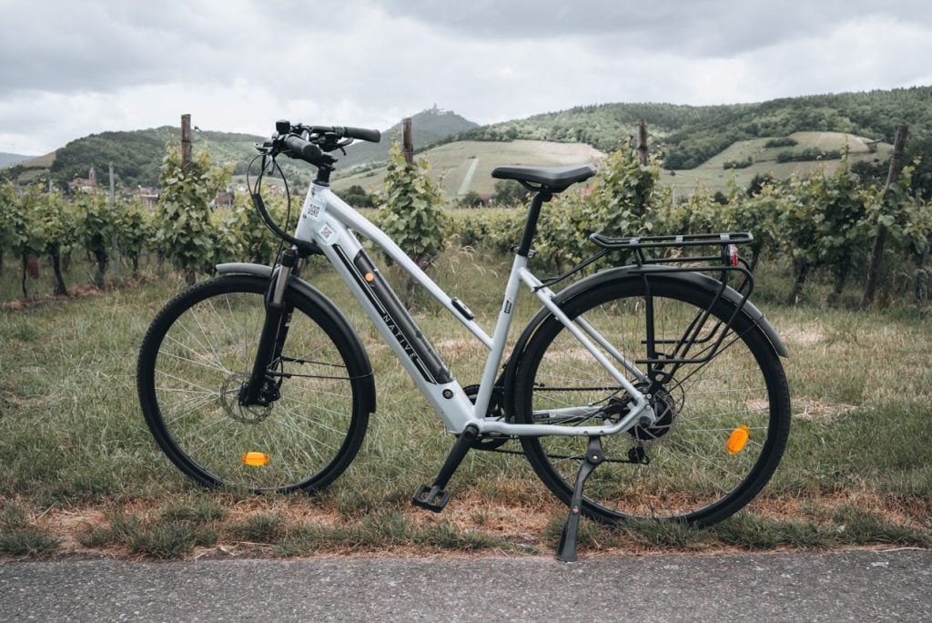 This is Jen Ciesielski's rental bike from Detour Alsace. It is an electrical bike that is parked next to some vineyards.