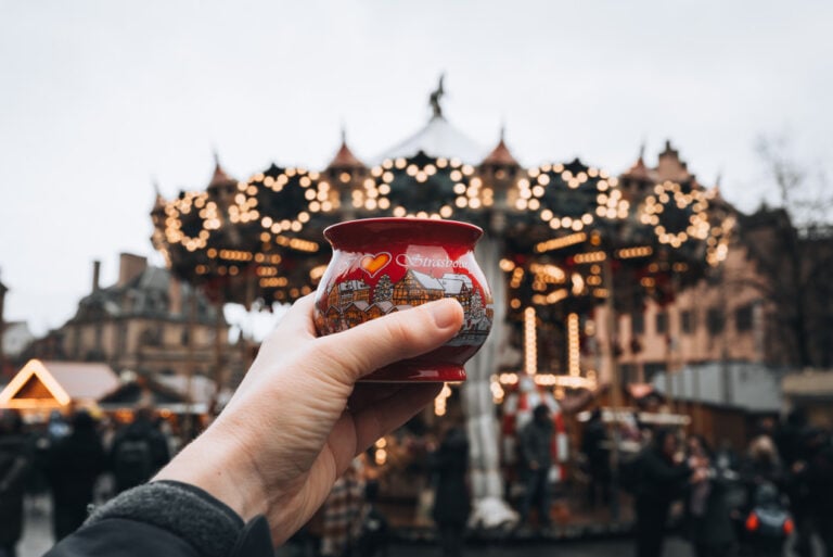 Jen holding a collectible Strasbourg Christmas market mug surrounded by festive lights and wooden market stalls. Sampling seasonal drinks at the Christmas markets is one of the most popular things to do this time of year.