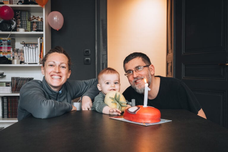 Jen with her son and husbands celebrating her son's first birthday on the weekend because they don't have to work. They are standing around a cake with a candle.