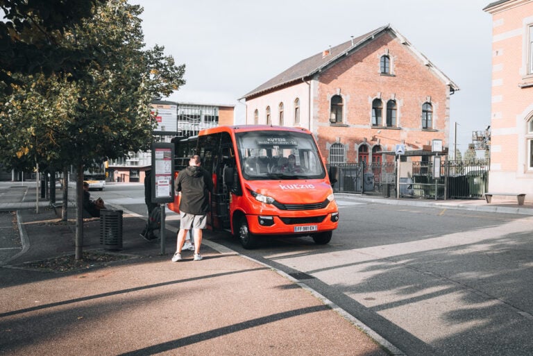 This is the Kutzig Bus at train station in Colmar. There is a sign with the name of the bus and a few people waiting to board.