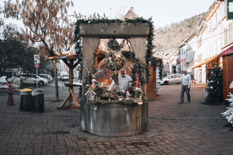 A local posing for a photo in front of the Fontaine des Vignerons at the Christmas market in Thann.