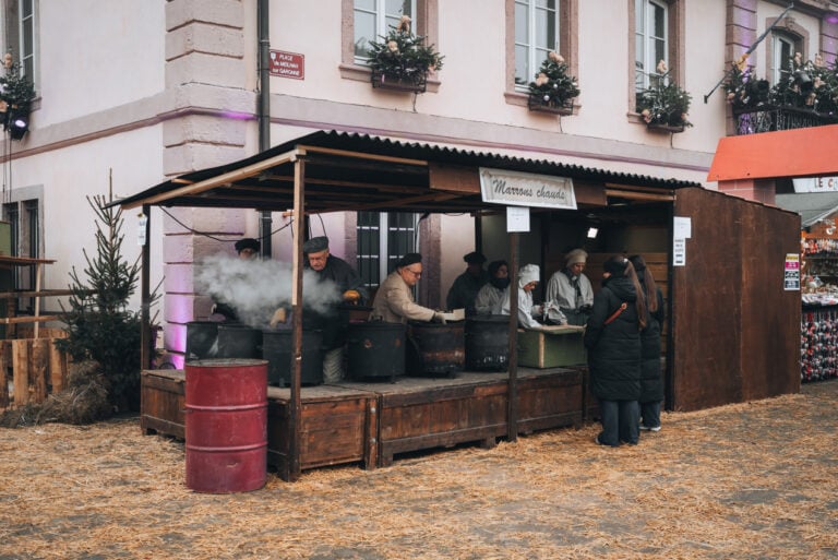 Locals roasting chestnuts in large round cast iron pots over a flame. Smoke is steaming out of the top and a small line of people has formed.