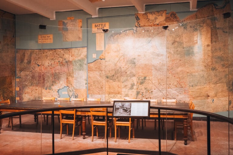 A rectangular table is in the center of a room surrounded by wooden chairs. On the walls there battle maps for World War 2. It was here that Germany surrendered and remains one of the significant historical sites in France.