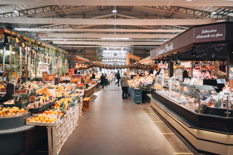 This is Marche Central de Nancy in Nancy. There is an aisle and one side there is fresh fruit and on the other a cheese vendor. In the distance people are walking around.