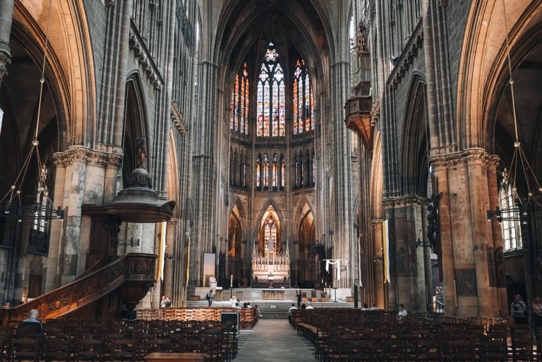 This is the interior of the Metz Cathedral. There is an aisle leading to the altar and wooden pews on either side. Above the altar there are stained glass windows.