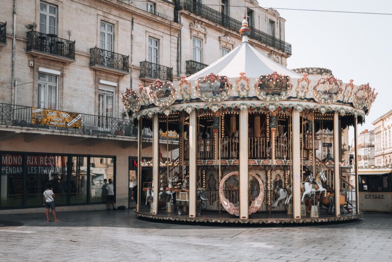 This is the the carousel in Place de la Comédie in Place de la Comédie. There are people on it, but it's stopped. The lights are also on. It's adjacent to a tall building. There are two people walking to the left of it.