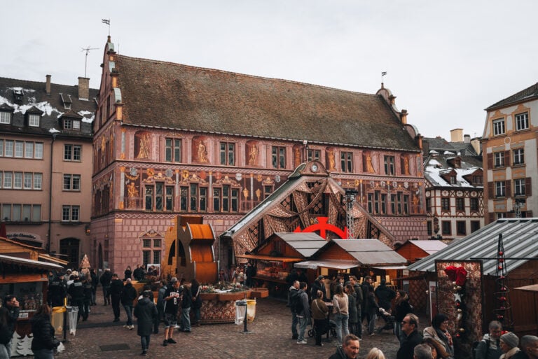 This Place de la Reunion during the Mulhouse Christmas market. There are wooden chalets, the history museum, and people walking around.