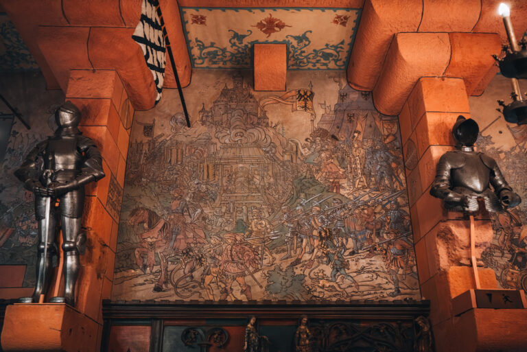 Giant mural showing a fighting scene from the Middle Ages in the Kaiser room of the Haut-Koenigsbourg Castle. On either side of the are armoured suits holding swords.