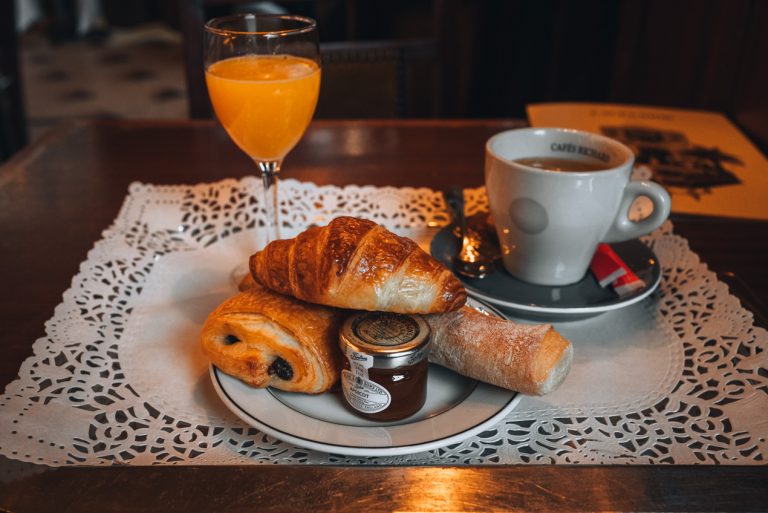 This is my breakfast from Brasserie Excelsior in Nancy. There is a plate with a jar of jam, one croissant, a piece of bread, and a pain au chocolate. Then, behind the dish, there is a glass of orange juice and a cup of coffee.