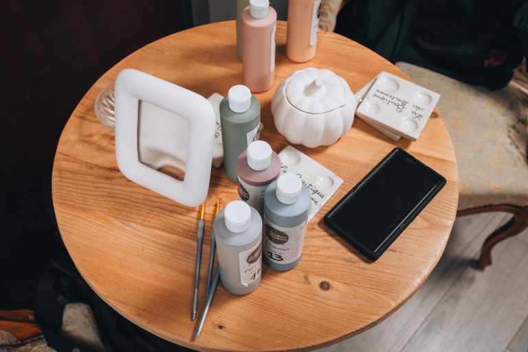 This is my picture frame before I started painting at La Boutique des Bouleaux. There is a picture frame and five bottles of paint and paint brushes on a round wooden table.