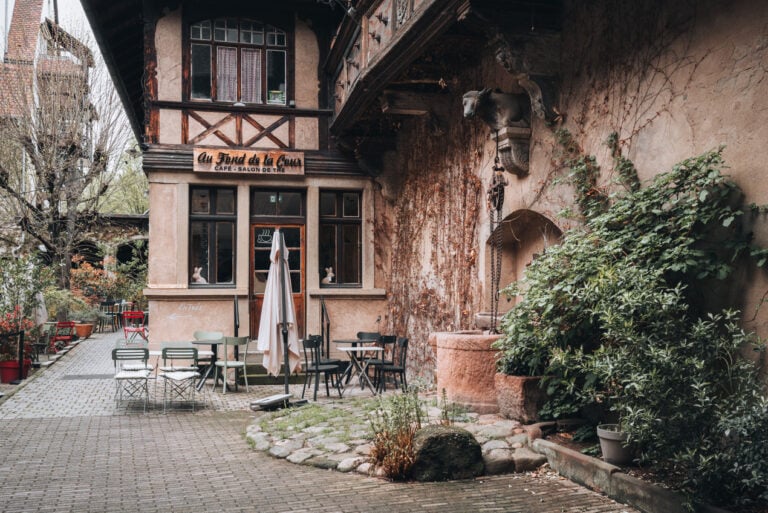 Quiet courtyard seating area at Au Fond de la Cour with café tables and chairs arranged around a small decorative fountain surrounded by greenery. The peaceful setting makes it a relaxing place to take a break while visiting Obernai.