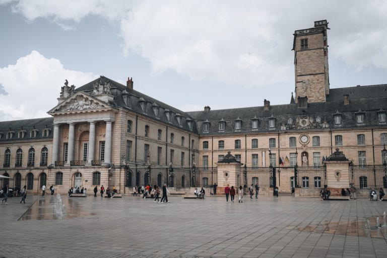 This is a panoramic view of the Palais des Ducs de Bourgogne in Dijon. It's a large stone palace with a tower and people are walking in front of it.
