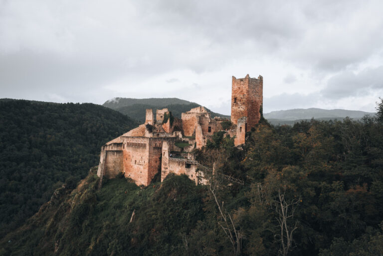Panorama of the Château de Saint-Ulrich along the Three Castles of Ribeauvillé Hike. It's a red stone castle surrounded by tree covered hill tops in the Vosges.