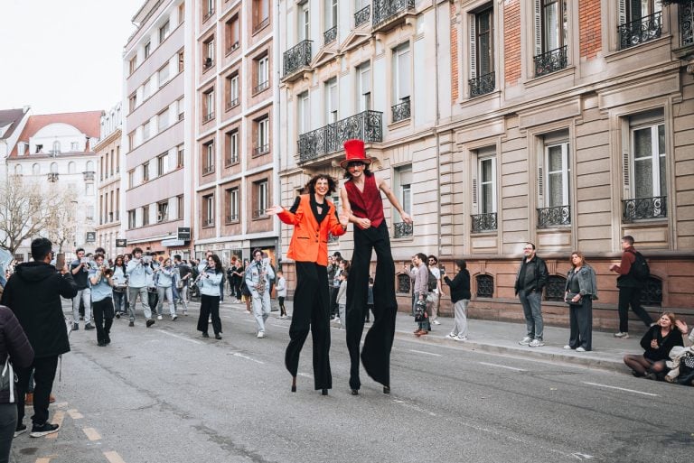 This is the parade at the carnival in Strasbourg. In the front their are two people on slits with make up and a band is walking behind them.
