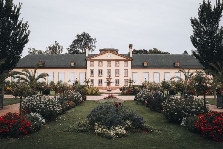 This is the Fontaine de l'Orangerie in Parc de l’Orangerie in Strasbourg. It's a large fountain surrounded by a grassed area and flower beds. In the background there is a stone building.