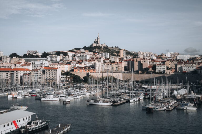 This is the view of Notre Dame de la Garde from the Passerelle Parvis-St. Jean. There is the church on the top of a hill in the distance and in the foreground there is the harbor filled with boats.