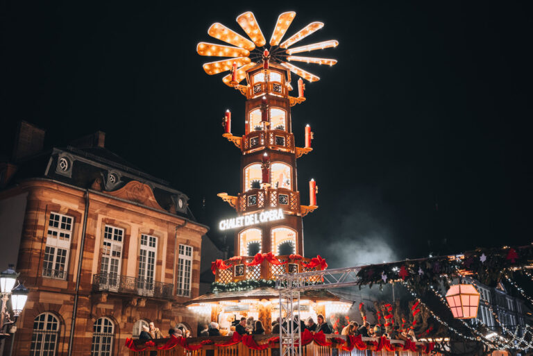 A wooden chalet with a tower at the Christmas market in Strasbourg's Place Broglie. It's a night so the lights are on and people are sitting at the bar on the top level.